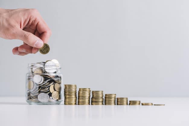 Investimentos com Maior rentabilidade - Mão de uma pessoal retirando uma moeda de um pote ao lado de uma pilha crescente de moedas