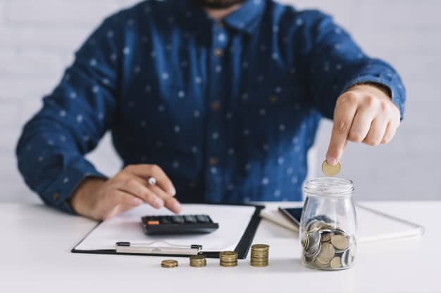 Investimento com maior rentabilidade: homem com uma das mãos usando uma calculadora e outra colocando uma moeda num jarro. na sua frente uma pilha crescente de moedas.